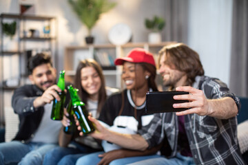 Blur background of four multi ethnic friends toasting with beer while talking selfie on modern smartphone. Focus on male hands holding cell phone. Young fans watching soccer match at home.