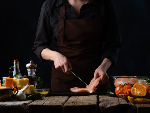 The photo shows the process of cooking poultry steaks. The cook cuts the meat for stuffing. Contrast colors - bright colors of ingredients and dark background.