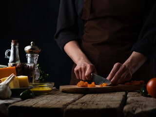 The cook prepares a vegetable salad. The photo shows the process of cutting vegetables for salad. On the left in the photo is cheese, sauce, spices. Home kitchen. Vitamins. Dark background.