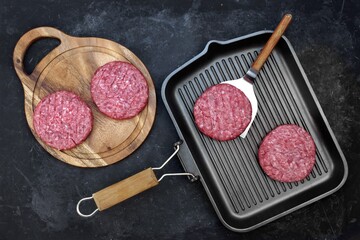 Griddle Grill Pap and Ground Beef Meat Patties for Grilling on Black Background, Overhead View. Raw Steak Burgers Cutlets On Grill Pan.  Burgers for Frying, Grill Tools and Cutting Board, Top View.