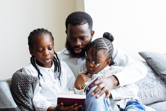 Father reads a book to his two daughters on sofa