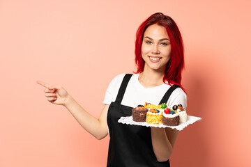 Pastry chef holding a muffins isolated on pink background pointing finger to the side