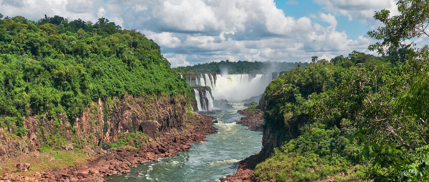 Iguazu waterfalls in Argentina, view from above. Panoramic view of many majestic powerful water cascades with mist. Panoramic image with reflection of blue sky with clouds.