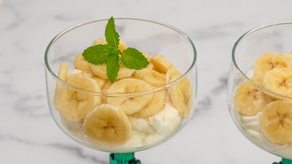 Whipped cream cheese dessert with sliced bananas and mint leaves served in glass dessert cups close up  on marble background with copy space