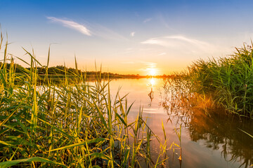 Scenic view at beautiful spring sunset with reflection on a shiny lake with green reeds, grass,...