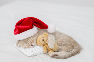 Cute kitten wearing red santa's hat sleeps under white blanket and hugs favorite toy bear