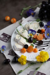 Camembert on a white plate with grape slices, physalis and viola flowers. A light snack in a light dish on a dark table.