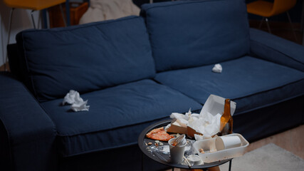 Chaos in empty living room with food garbage, bottle of beer and napkins on blue sofa. Unorganized house apartment of alone woman with sever depresion having trash, rubbish with no people in