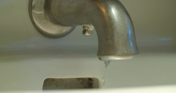 Slow motion medium shot of a faucet as the water shuts off and drips of water continue to fall.
