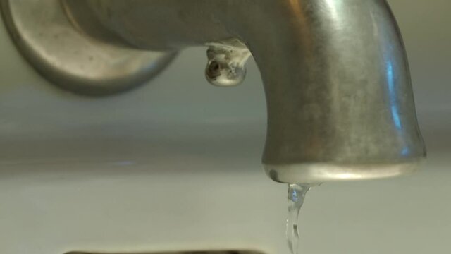 Slow motion medium shot of a faucet as the water runs, shuts off and drops of water continue to drop.