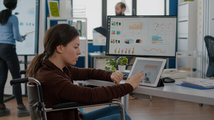 Multitasking paralyzed project manager, businesswoman with disabilites invalid immobilized woman using computer and tablet in same time working in business start-up office sitting in wheelchair.