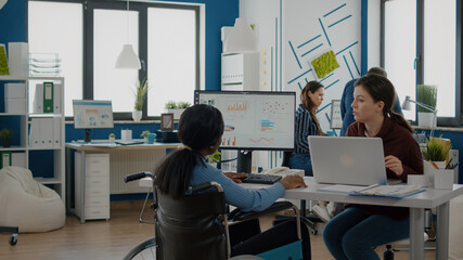 African paralysed invalid immobilized woman in wheelchair and colleague analyzing datas laptop while sitting in start up business office. Colleagues working in background searching financial solutions