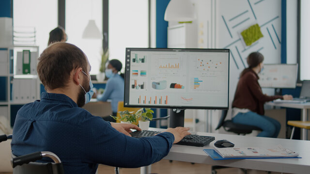 Immobilized business man with protective mask working in new normal business financial company typing on pc, checking reports, analysing data looking at desktop sitting in wheelchair