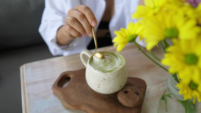 Hand stirring matcha tea latte the spoon and shows the foam to the camera. Yellow flowers on the table