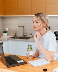 Portrait of a beautiful woman working remotely on a laptop, blonde in a white t-shirt at the table in the kitchen with a laptop