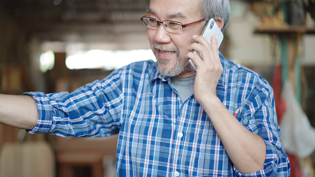 Asian senior male entrepreneur or carpenter talking on the phone with customer in carpentry workshop. happy feeling for success dealing.