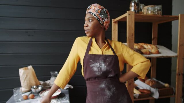 Zoom in shot of beautiful afro-american female baker in apron standing with hand on heap in kitchen, looking at camera and smiling
