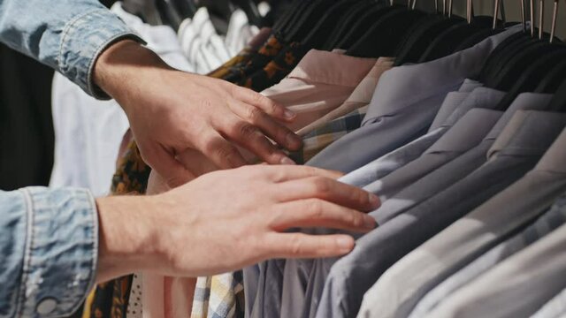 Close-up of unrecognizable male hands sorting shirts of different style and colors hanging on rails in modern clothing store
