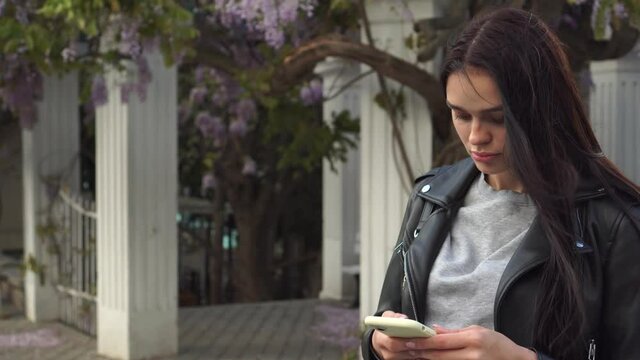 Smiling attractive beautiful cute young brunette girl in a leather jacket uses a smartphone. Flowering wisteria vine in the city's spring park on the background