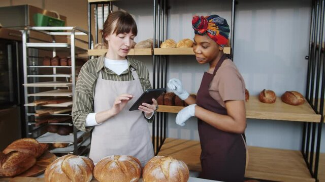 Young Caucasian woman using digital tablet and speaking with Afro-American female colleague as she putting fresh bread on wooden display rack during workday in bakery