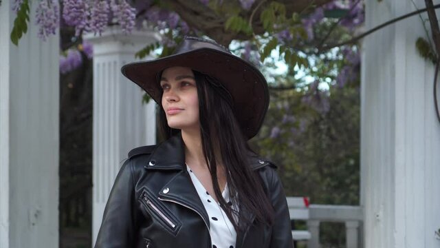 Portrait of a young attractive stylish brunette woman in a hat, black leather jacket. A model on the background of a flowering wisteria vine in the city's spring park