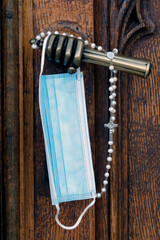 Old metal church door handle. Blue Surgical mask and Rosary.  Covid-19 epidemic.