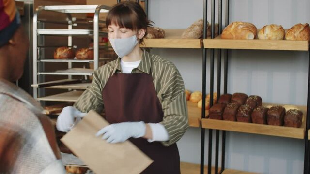 Over the shoulder shot of Caucasian saleswoman in mask and gloves putting loafs of bread in paper bag for black female customer while working in bakery during covid-19 pandemic