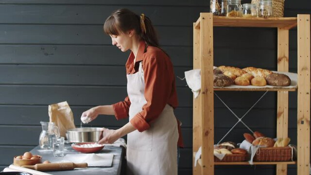 Young Caucasian woman in apron using sifting flour on table with steel sieve while working in bakery