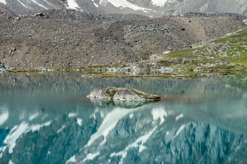 Rock in turquoise mountain lake. Snowy mountain reflected in azure clear water of glacial lake. Beautiful sunny background with snow-white glacier reflection in green water surface of mountain lake.