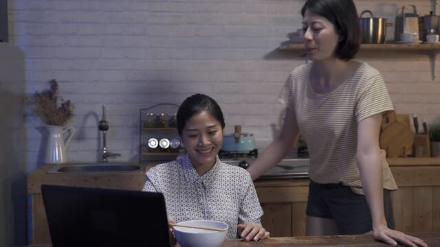 considerate lesbian is carrying a bowl of noodles over and standing behind showing concern as her girlfriend is working overtime on laptop at home.