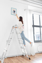 home improvement, decoration and people concept - happy smiling woman on ladder decorating home with art © Syda Productions