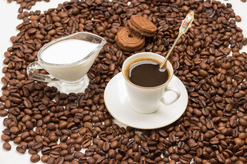 Cup of coffee, cream and cookies among roasted coffee beans