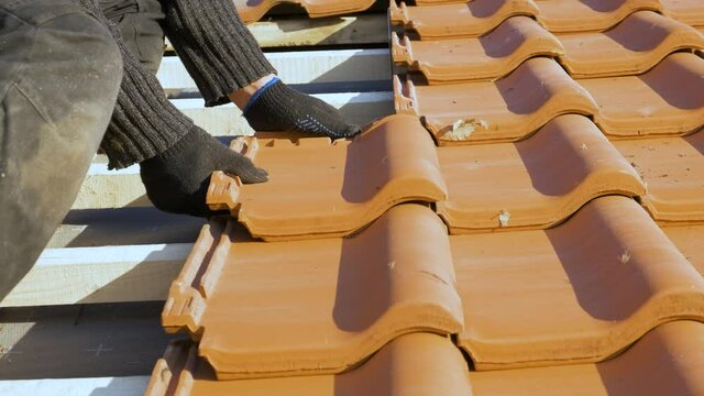 Closeup of worker hands installing yellow ceramic roofing tiles mounted on wooden boards covering residential building roof under construction.