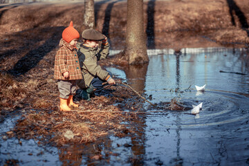 Cute little boy with his baby sister in funny hat are playing with paper boats in the spring park. Image with selective focus and toning