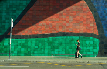 chica joven de pelo corto y rojo camiando con la maleta por el aeropuerto de Barcelona hacia la parada del autobus mirando el movil con un mosaico gigante detras  - obrazy, fototapety, plakaty