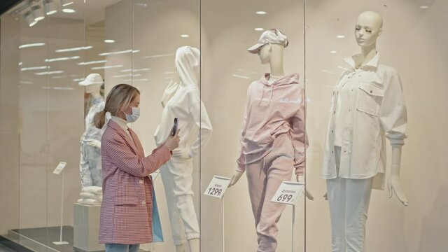 Medium shot of young Caucasian woman in face mask standing at window display of fashionable clothing store taking photo of outfit on mannequin on smartphone