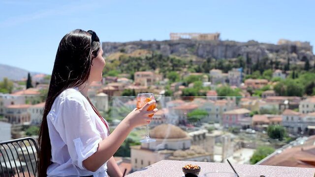 A beautiful tourist woman enjoys a drink on a rooftop bar overlooking the old town and Acropolis of Athens, Greece, during her summer holiday time