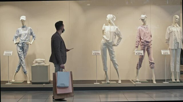 Full stab shot of stylish handsome man in face mask passing by window displays of stylish clothing store standing in front of mannequin to take picture of its outfit on smartphone