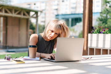 Beautiful serious thinking woman working at laptop outside and drinking coffee. Using computer and...