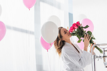 Young woman in bathrobe smelling roses near balloons in bathroom - obrazy, fototapety, plakaty