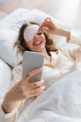 Obraz na płótnie Canvas Smartphone in hand of blurred woman in sleeping mask on bed