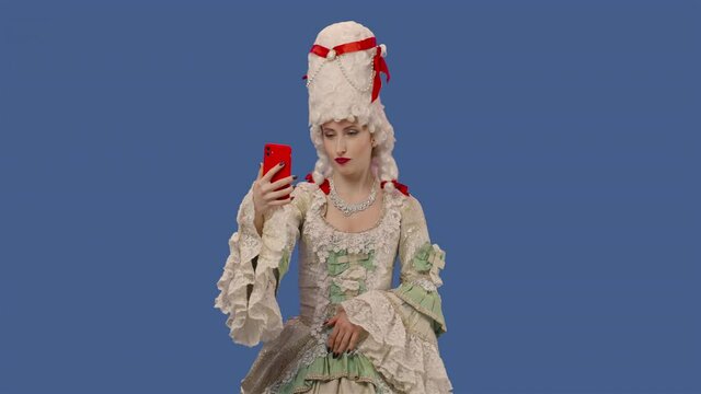 Portrait of courtier lady in white vintage lace dress and wig talking for video chat using mobile phone. Young woman posing in studio with blue screen background. Close up. Slow motion ready 59.94fps.