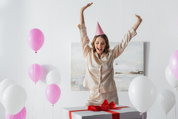 Positive woman in pajama standing near huge present and balloons at home