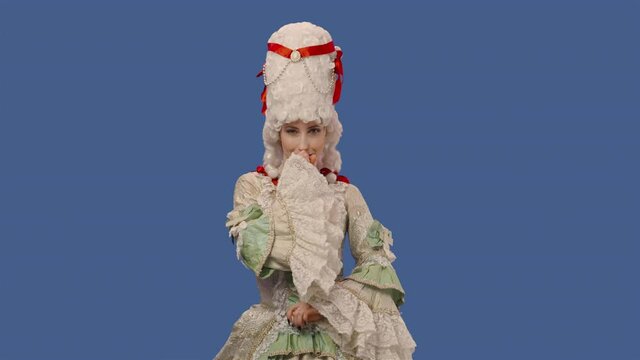 Portrait of courtier lady in white vintage dress and wig smiling coquettishly, making eyes, winking. Young woman posing in studio with blue screen background. Close up. Slow motion ready 59.94fps.