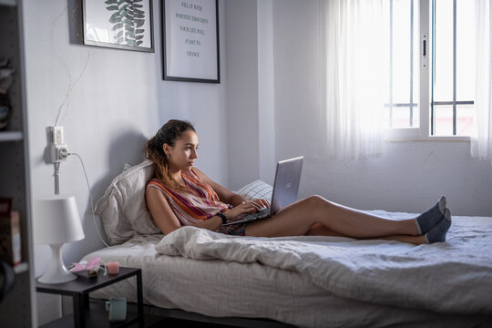 Young woman with dark hair on the bed using the computer. 
young woman working from his house on the bed with light coming from thw window