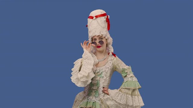 Portrait of courtier lady in white vintage lace dress, wig and sunglasses smiles coquettishly and flirts. Young woman posing in studio with blue screen background. Close up. Slow motion ready 59.94fps
