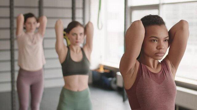 Waist-up of young Mixed-Race woman doing triceps extensions with barbell weight along with blurred athletes standing in one row behind her in gym