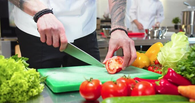 Male tattooed hands cutting tomato. Man cook working in a busy restaurant kitchen, slicing vegetables, preparing salad dish. Close up shot. Workflow at cafeteria kitchen. Cookery, culinary profession