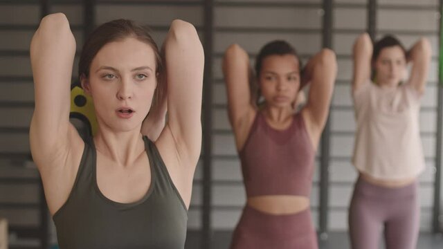 Waist-up of slim Caucasian woman doing triceps extensions with barbell weight along with blurred athletes standing behind her in gym