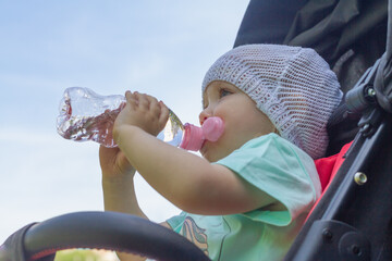 Baby girl drinks water from a plastic bottle in the summer in the heat sitting in a stroller